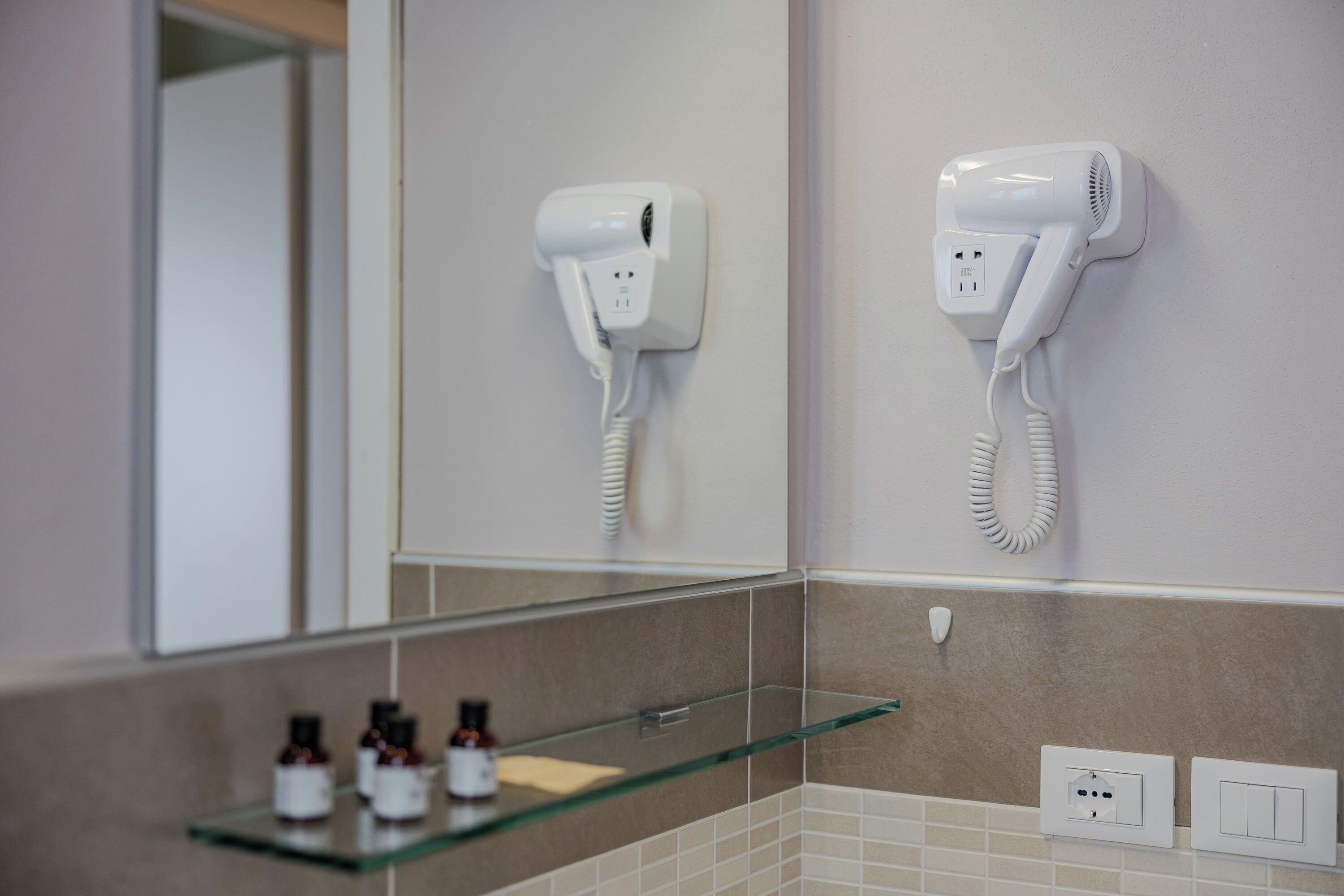 A bathroom with a hairdryer mounted on the wall, a glass shelf with toiletries, and a mirror.