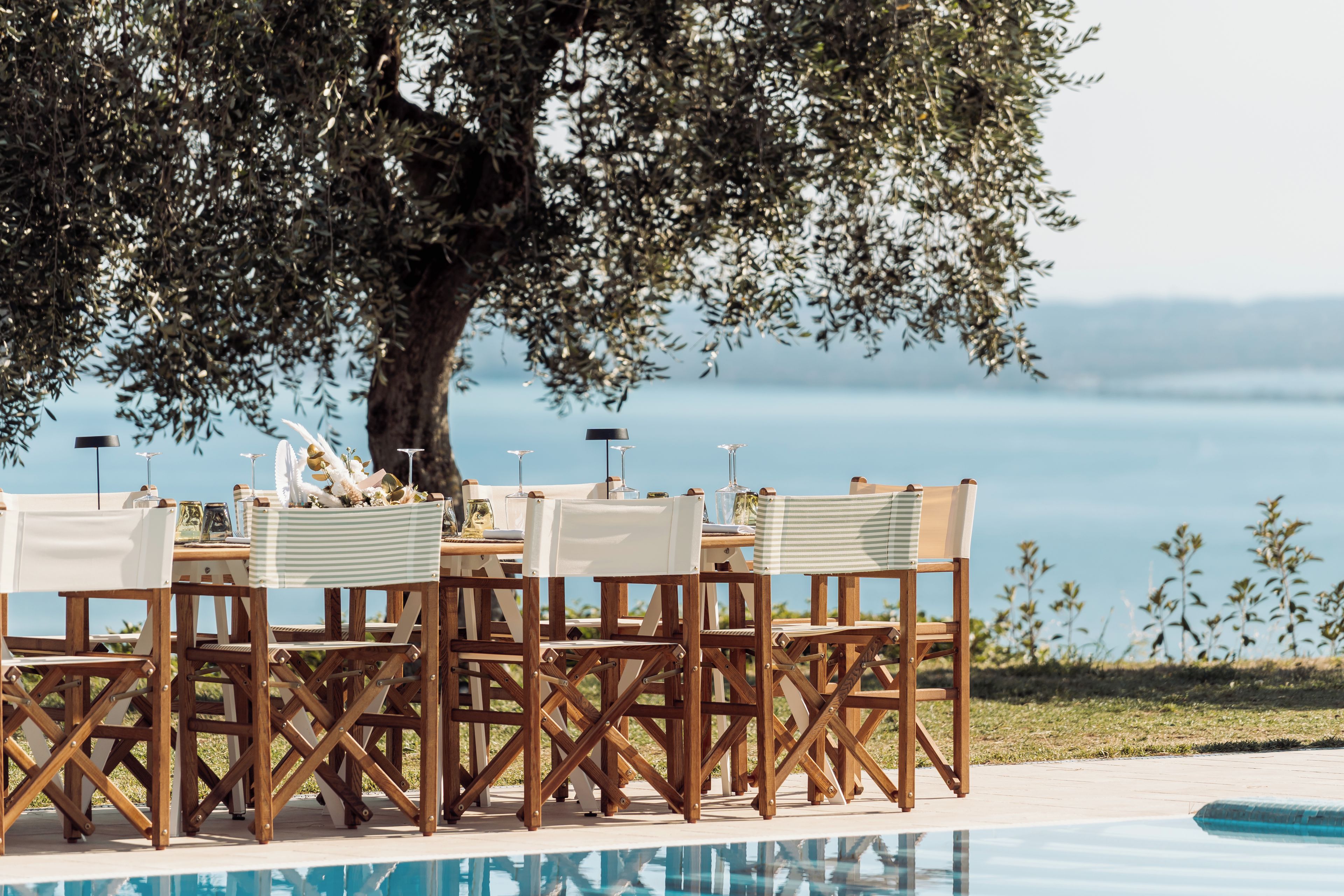 Área de refeições ao ar livre com piscina, cadeiras de madeira e mesa posta para uma refeição.