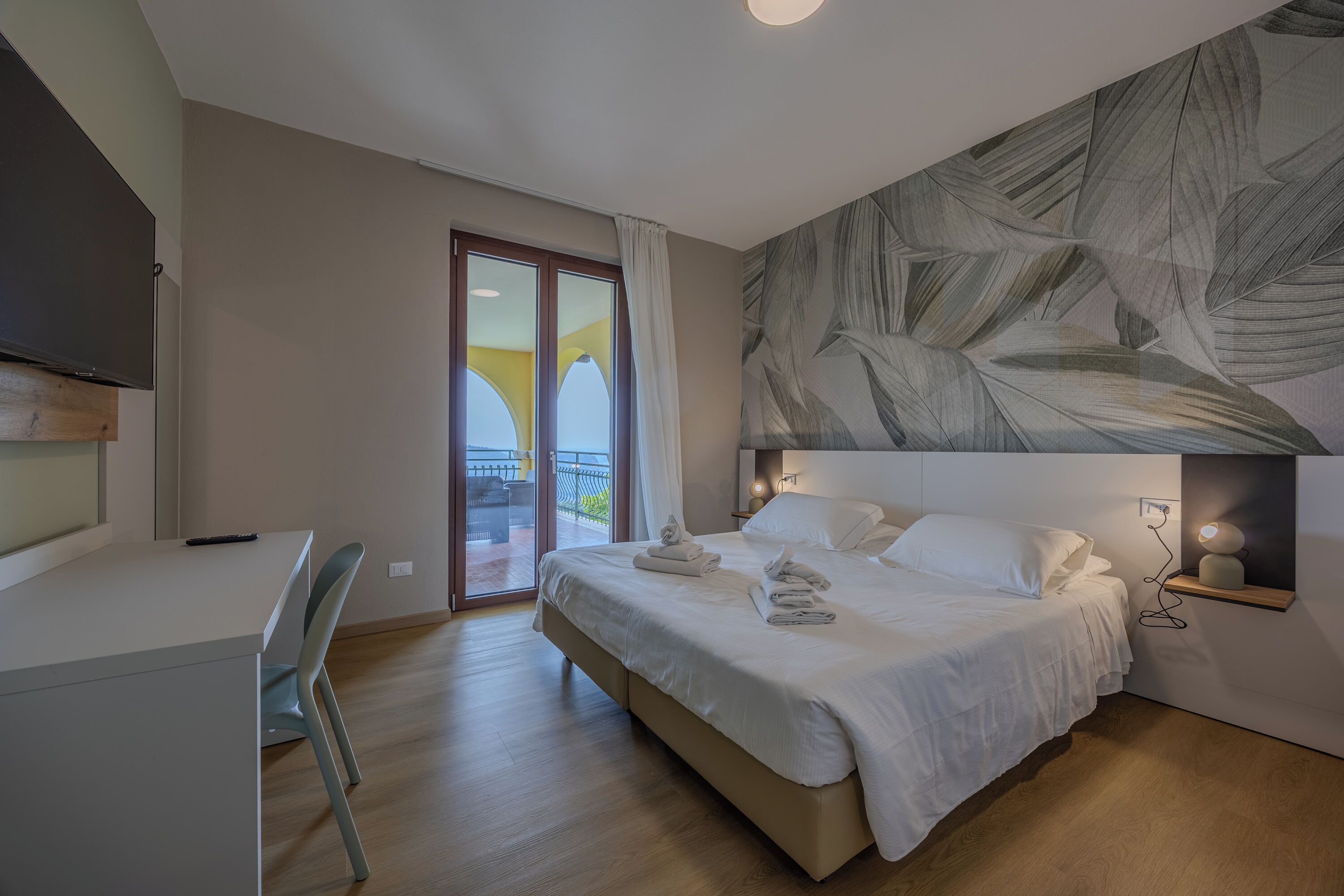Un dormitorio con una cama, un escritorio y una silla. Se ve una ventana con vistas a un balcón.