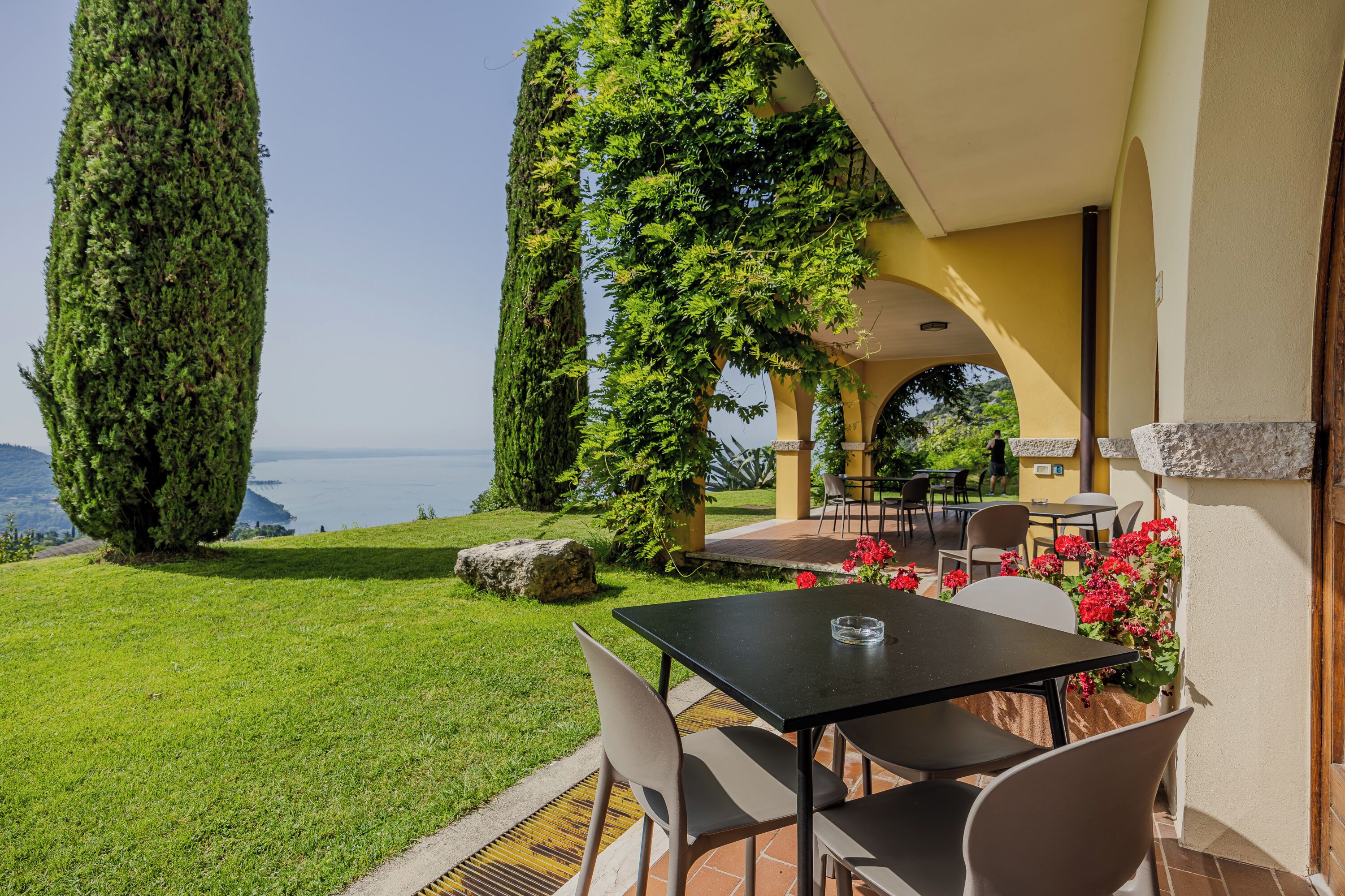 A patio with a table and chairs, surrounded by greenery and a view of the sea.