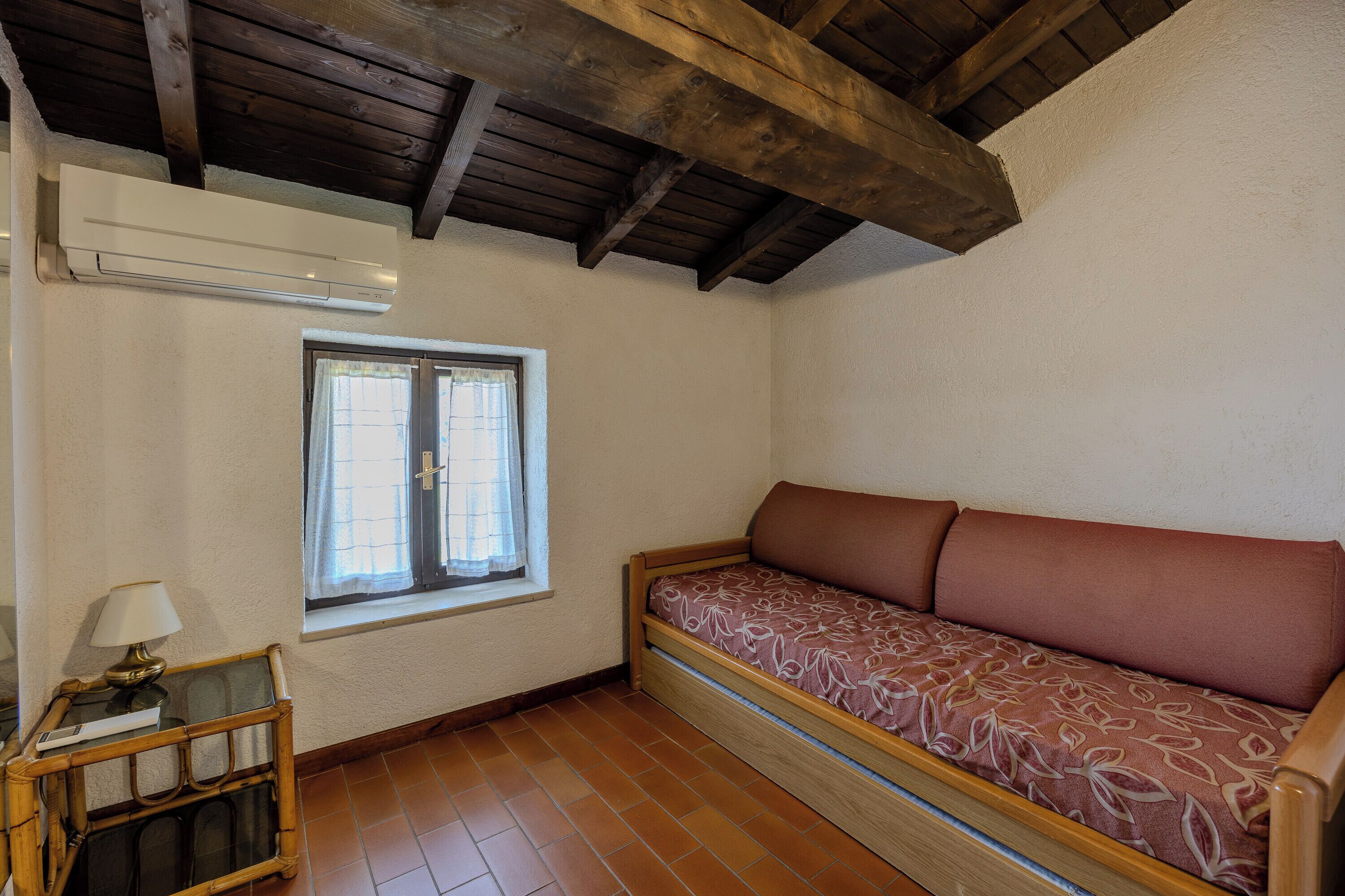 A room with a wooden ceiling, a window with curtains, a sofa with a patterned cover, and a small table with a lamp.