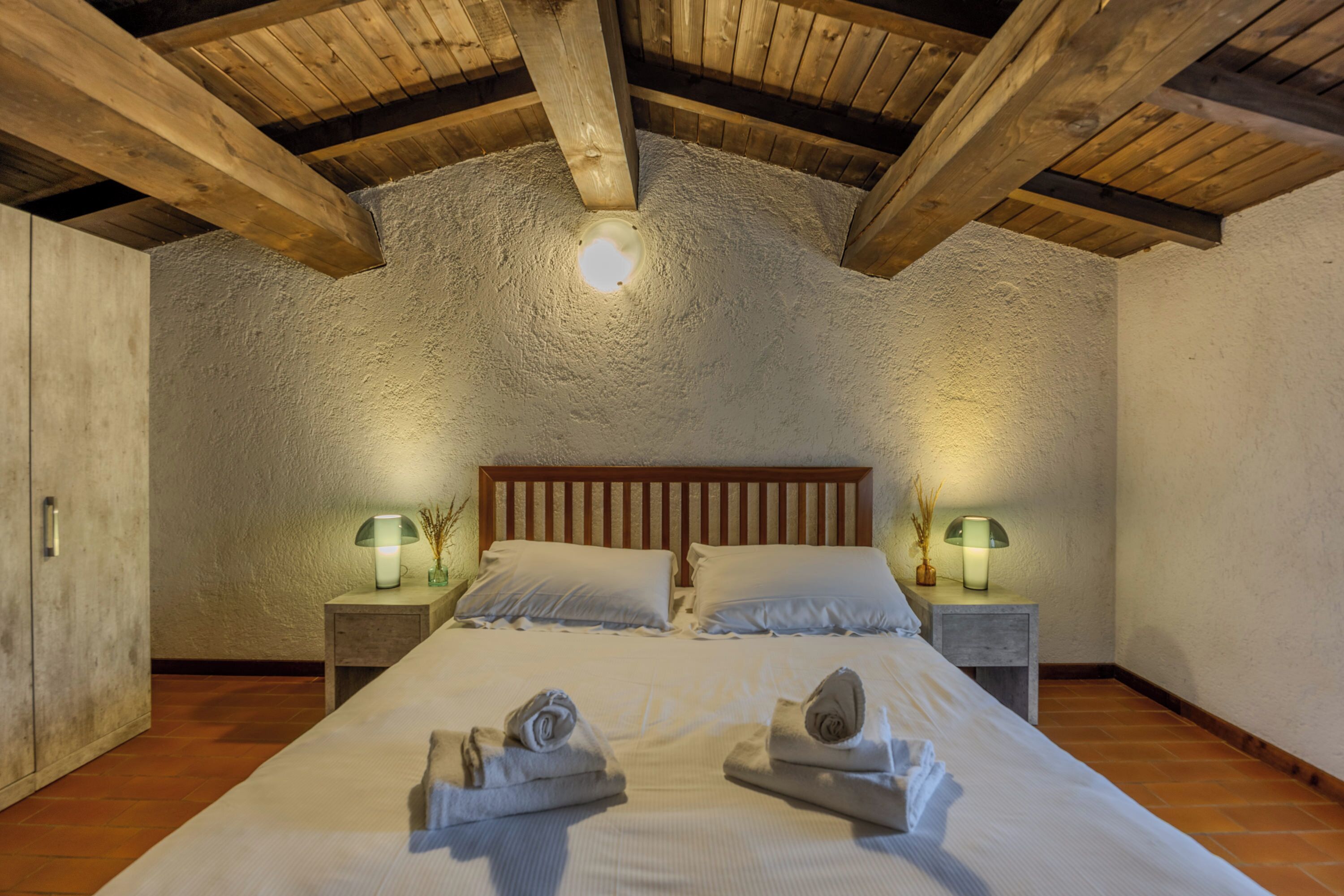 Un dormitorio con techo de madera, una cama grande con ropa blanca, dos mesitas de noche con lámparas y una mesita auxiliar con lámpara.