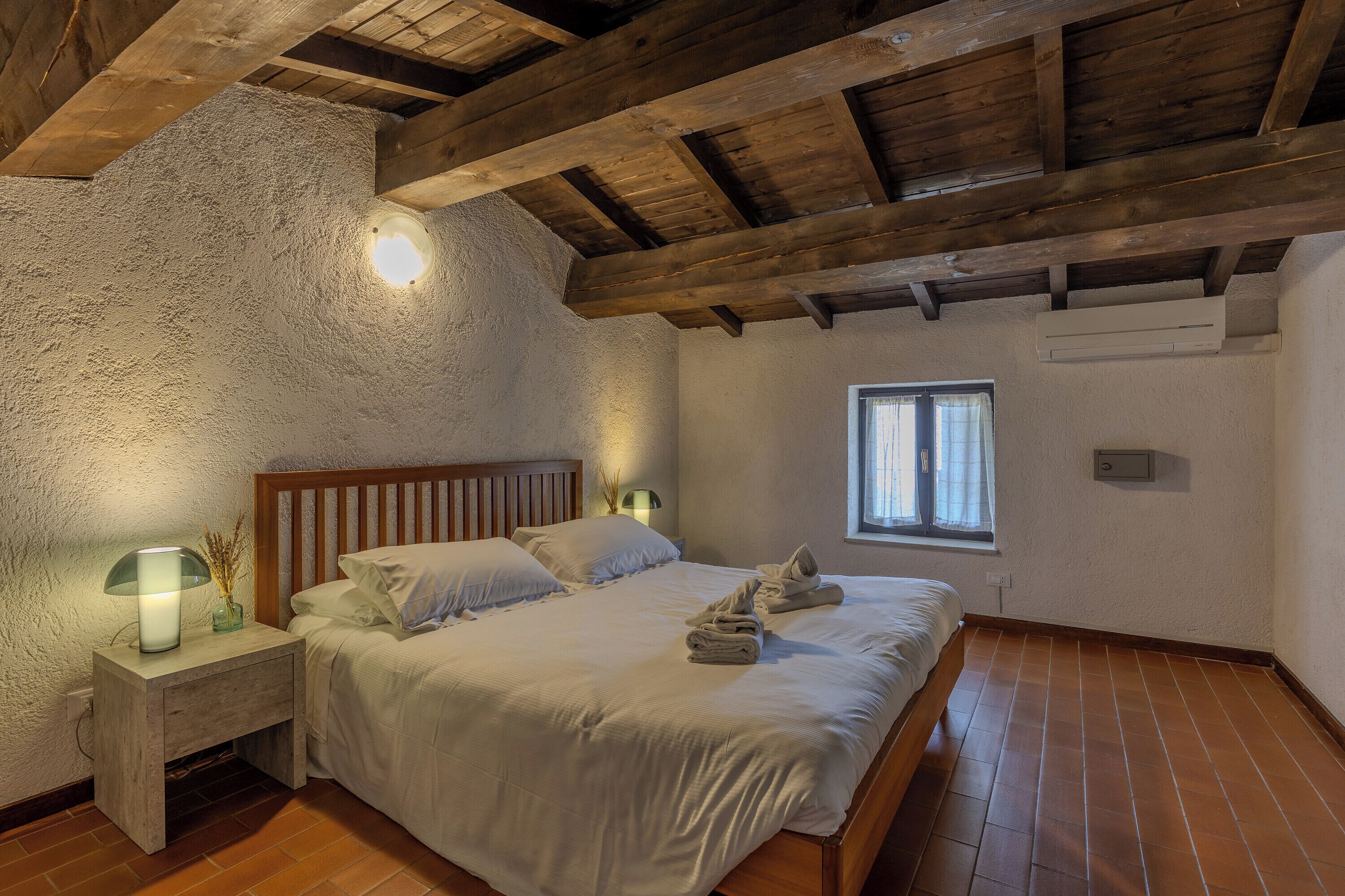 Una camera da letto con soffitto in legno, un letto con lenzuola bianche, un comodino con una lampada e una finestra vista esterna.