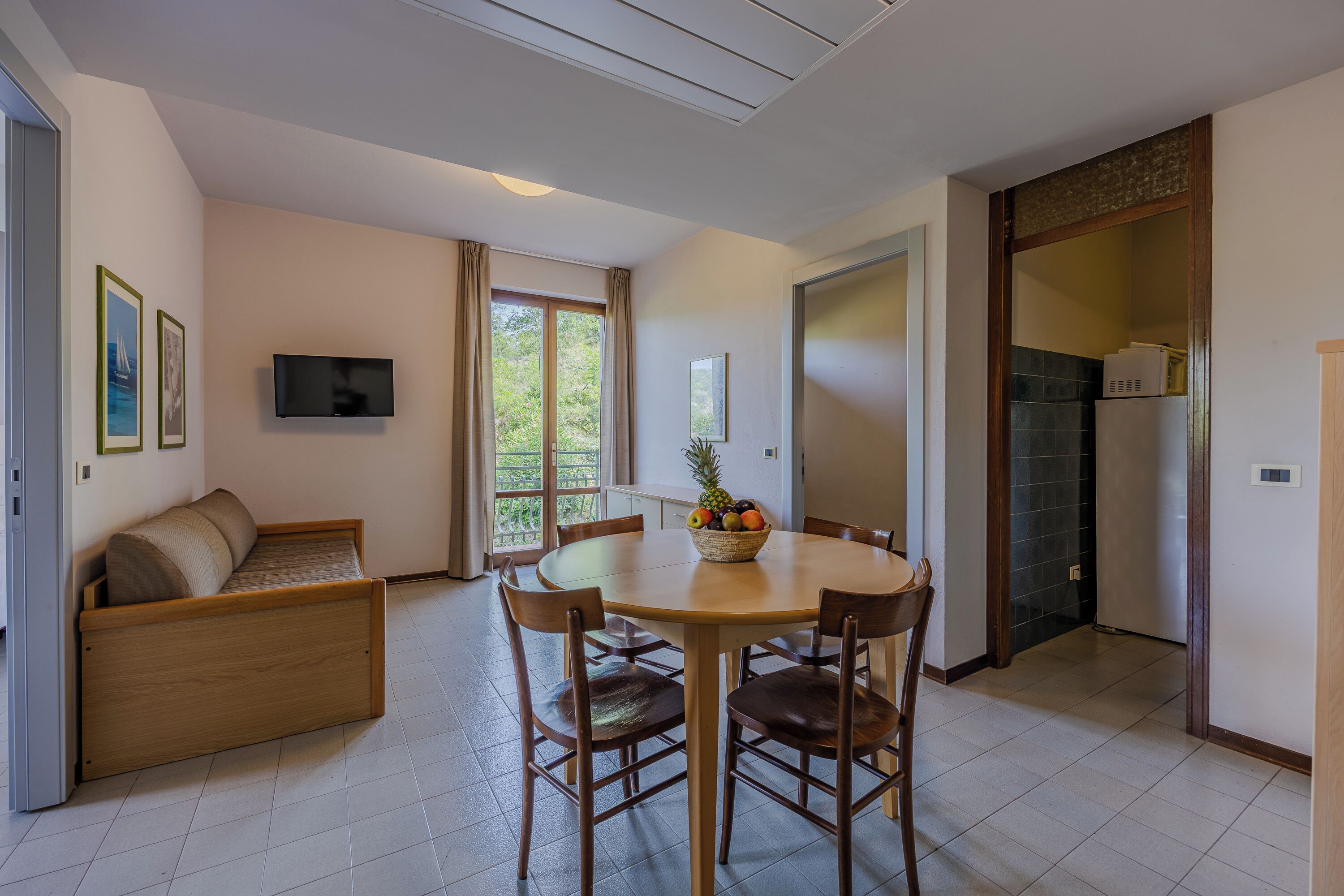 Una sala de estar moderna con un sofá, una mesa de comedor con sillas, un televisor de pantalla plana montado en la pared y un frutero sobre la mesa.