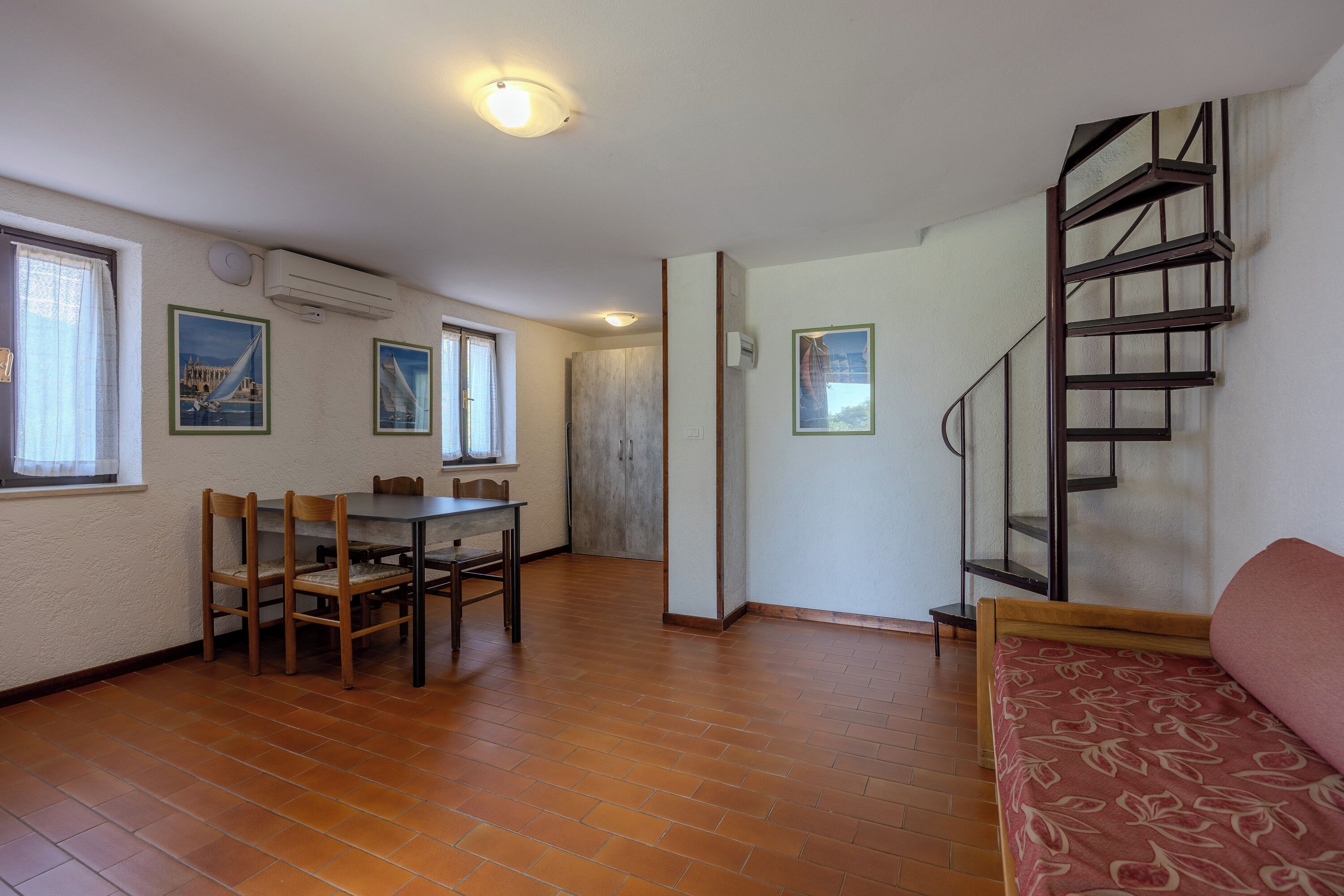 A room with a dining table and chairs, a staircase, and a bed with a patterned bedspread.