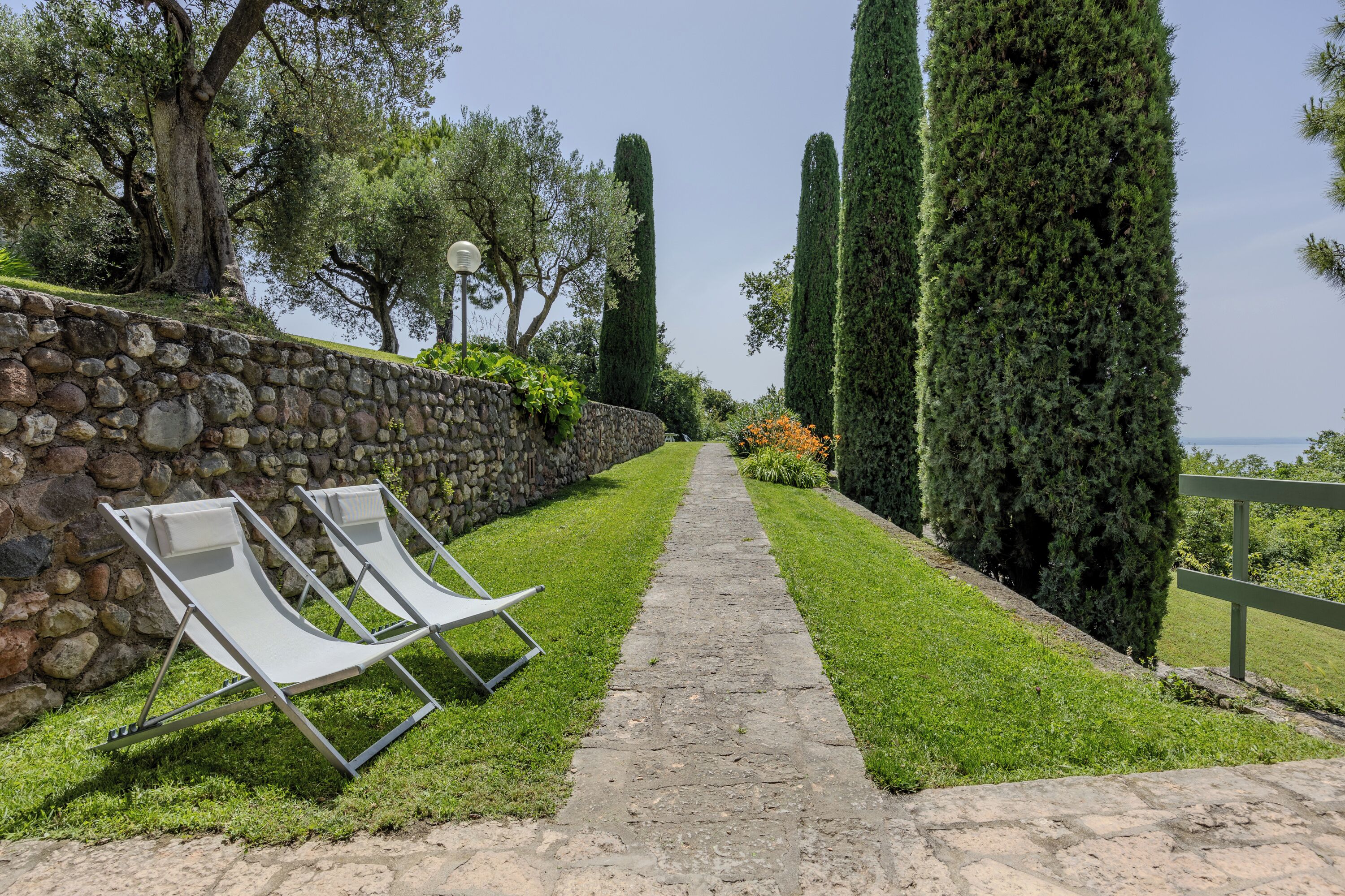 A stone wall with two lounge chairs and a pathway leading to a body of water.