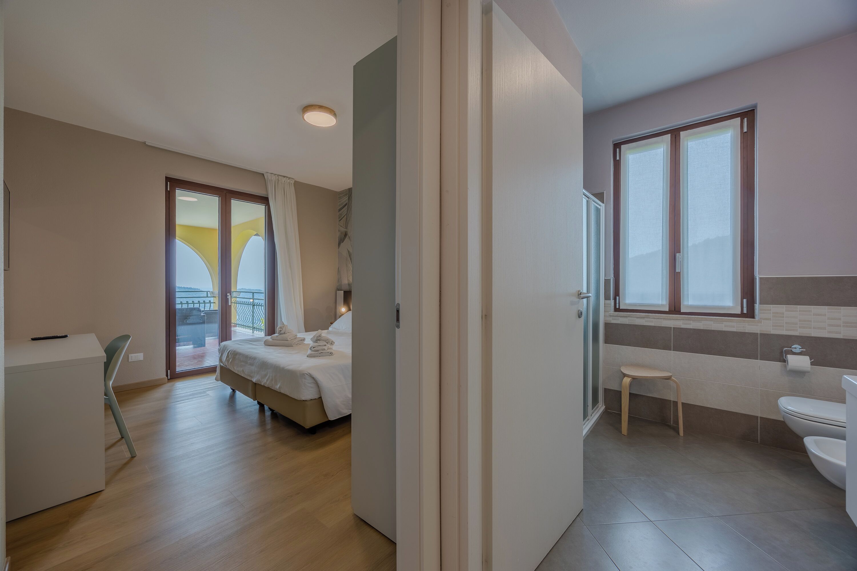 Habitación de hotel con cama, escritorio, silla, baño con inodoro y lavamanos, y balcón con vistas.