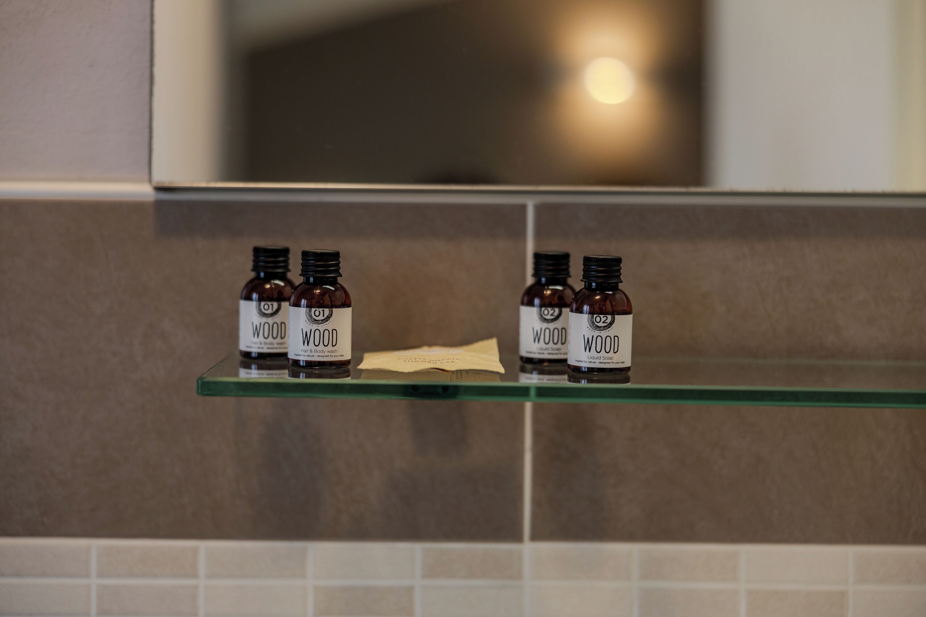 A glass shelf with four small bottles labeled "WOOD" and "Liquid Wood Body Wash" in a bathroom setting.
