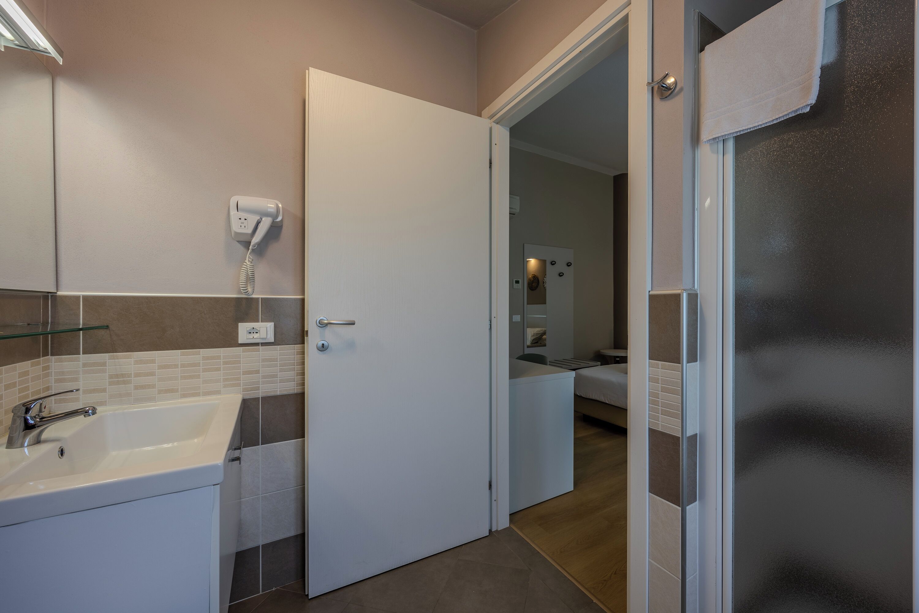 Un baño con un lavamanos blanco, un secador de pelo en la pared y una puerta que da a un dormitorio.