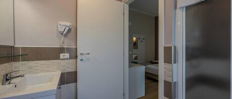 Un baño con un lavamanos blanco, un secador de pelo en la pared y una puerta que da a un dormitorio.