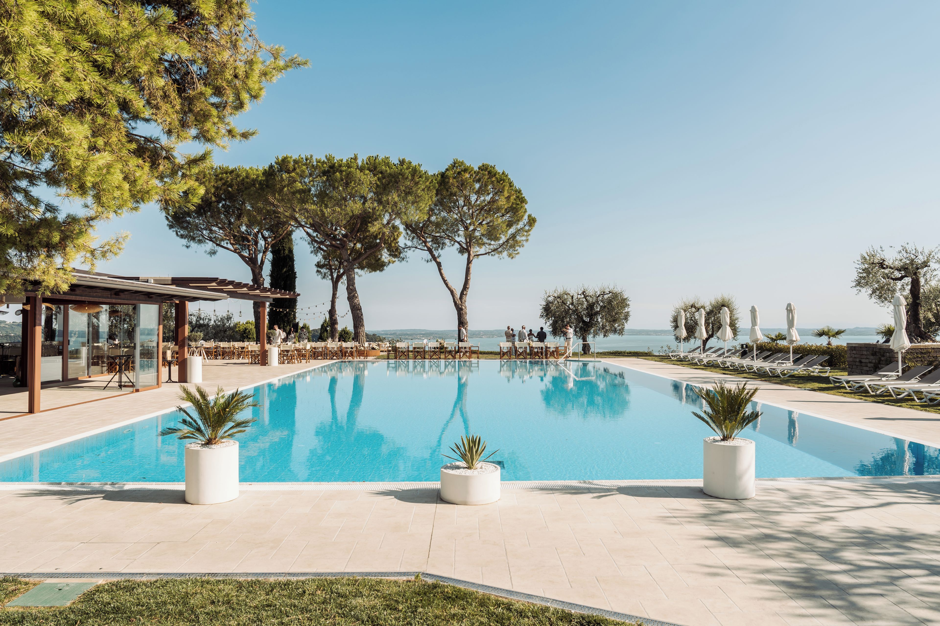 A clear blue swimming pool with lounge chairs and umbrellas, surrounded by trees and a view of the sea.