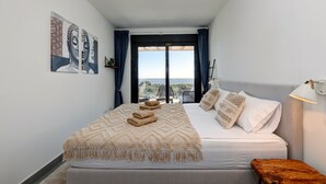 Une chambre à coucher avec un lit, des oreillers, une table de chevet, une lampe, une fenêtre avec des rideaux et des œuvres d’art accrochées au mur.