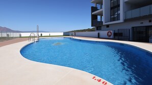 Une piscine avec un panneau indiquant la présence d’un sauveteur, un ciel dégagé et un immeuble doté de balcons.