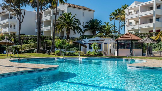 A swimming pool with a tiled edge, surrounded by palm trees and a building with balconies. - 2-Bed Apartment in Miraflores with Pool, Terrace, Garden & Wi-Fi (Mijas)