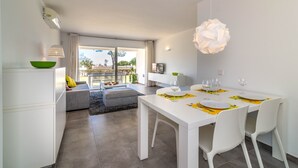 A modern dining area with a white table set for two, a green bowl on a white cabinet, and a view of the outdoors. - 2-Bed Apartment in Miraflores with Pool, Terrace, Garden & Wi-Fi (Mijas)