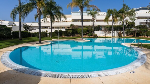 Ein Swimmingpool mit klarem, blauem Wasser, umgeben von Palmen und einem Gebäude mit mehreren Balkonen im Hintergrund.