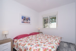 Een slaapkamer met een bed, een nachtkastje, een lamp, een raam en een schilderij aan de muur.