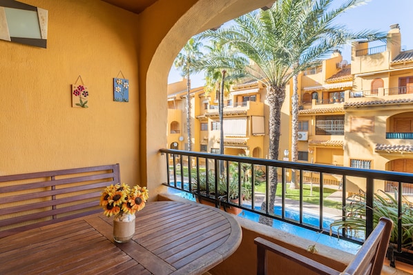 A balcony with a wooden table and chairs, a vase of sunflowers, and a view of a swimming pool and palm trees.