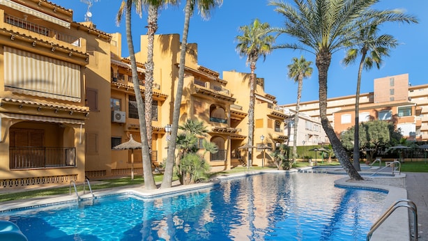 A swimming pool with a clear blue water surface, surrounded by palm trees and buildings with balconies and air conditioning units. - Apartment "El Albero" with Pool, Terrace & Wi-Fi (La Mata Torrevieja Alicante)
