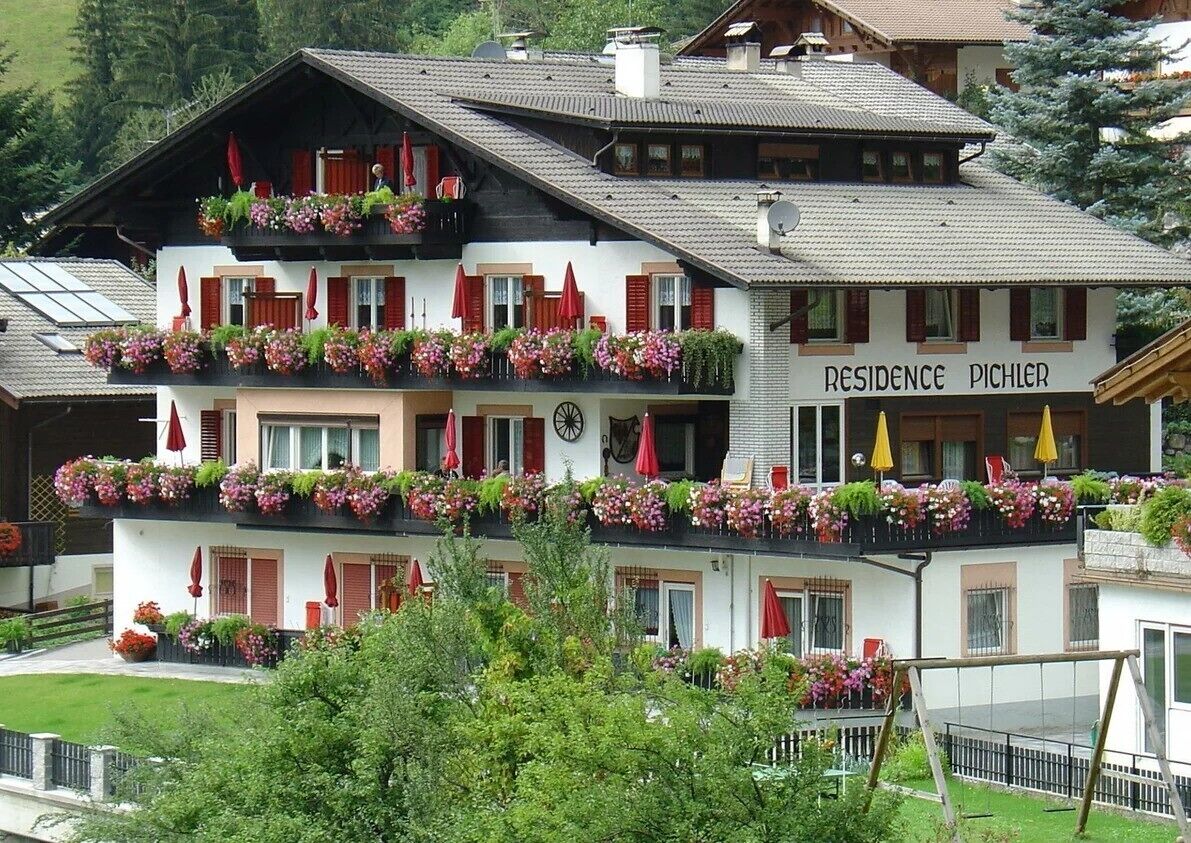 Un immeuble à plusieurs étages, doté de balcons ornés de fleurs, une enseigne indiquant « RÉSIDENCE PICHLER », et une piste de ski visible au loin.