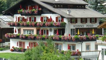 Un immeuble à plusieurs étages, doté de balcons ornés de fleurs, une enseigne indiquant « RÉSIDENCE PICHLER », et une piste de ski visible au loin.