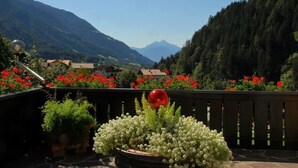 Ein Balkon mit Topfpflanzen, einer roten, kugelförmigen Gestalt und Blick auf eine gebirgige Landschaft.