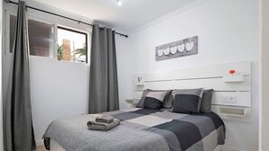 Een slaapkamer met een bed, gordijnen, een nachtkastje, een wandplank en een raam met uitzicht.