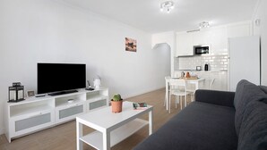 Een moderne woonkamer met een flatscreen televisie, een witte salontafel, een grijze bank en een keuken met witte kasten en apparatuur.