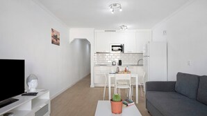 Een moderne woonkamer met een flatscreen-tv, een grijze bank, een witte salontafel met een potplant en een keuken op de achtergrond.