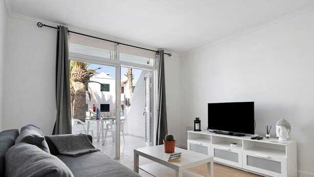 Een moderne woonkamer met een grijze bank, een witte salontafel, een flatscreen televisie op een wit tv-meubel, en een schuifdeur die leidt naar een buitenruimte.
