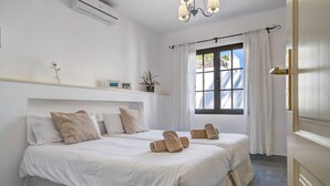 A hotel room with two beds, a window with curtains, a mounted air conditioner, a chandelier, and a small shelf with decorative items. - Villa 'Princesa Faina' with Partial Sea Views, Private Pool and Wi-Fi (Tias)