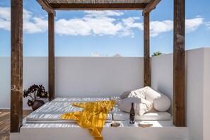 Un espace extérieur ensoleillé, aménagé avec une pergola en bois, un banc blanc doté de coussins et un pot de fleurs.