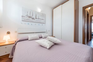 A bedroom with a bed, a nightstand with a lamp, a wall-mounted painting, and a door leading to another room.