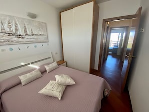 A bedroom with a bed, pillows, a wardrobe, a painting, and a hallway leading to another room.
