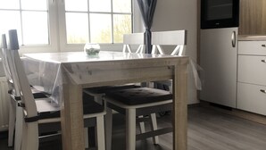 En matplats med ett träbord och stolar, ett fönster med gardiner och ett köksområde med kylskåp och ugn.