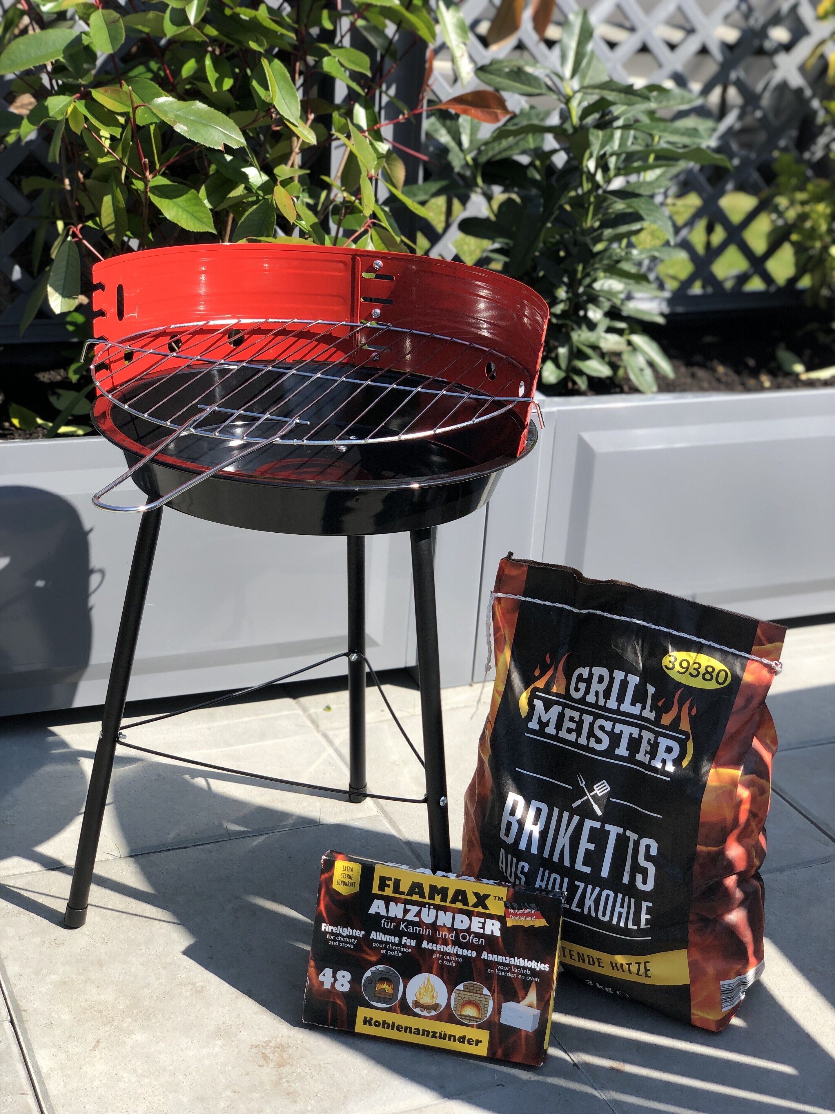 A red barbecue grill with a metal grate, a bag of charcoal labeled 'Flamax', and a box of charcoal labeled 'Kohlenanzünder'.