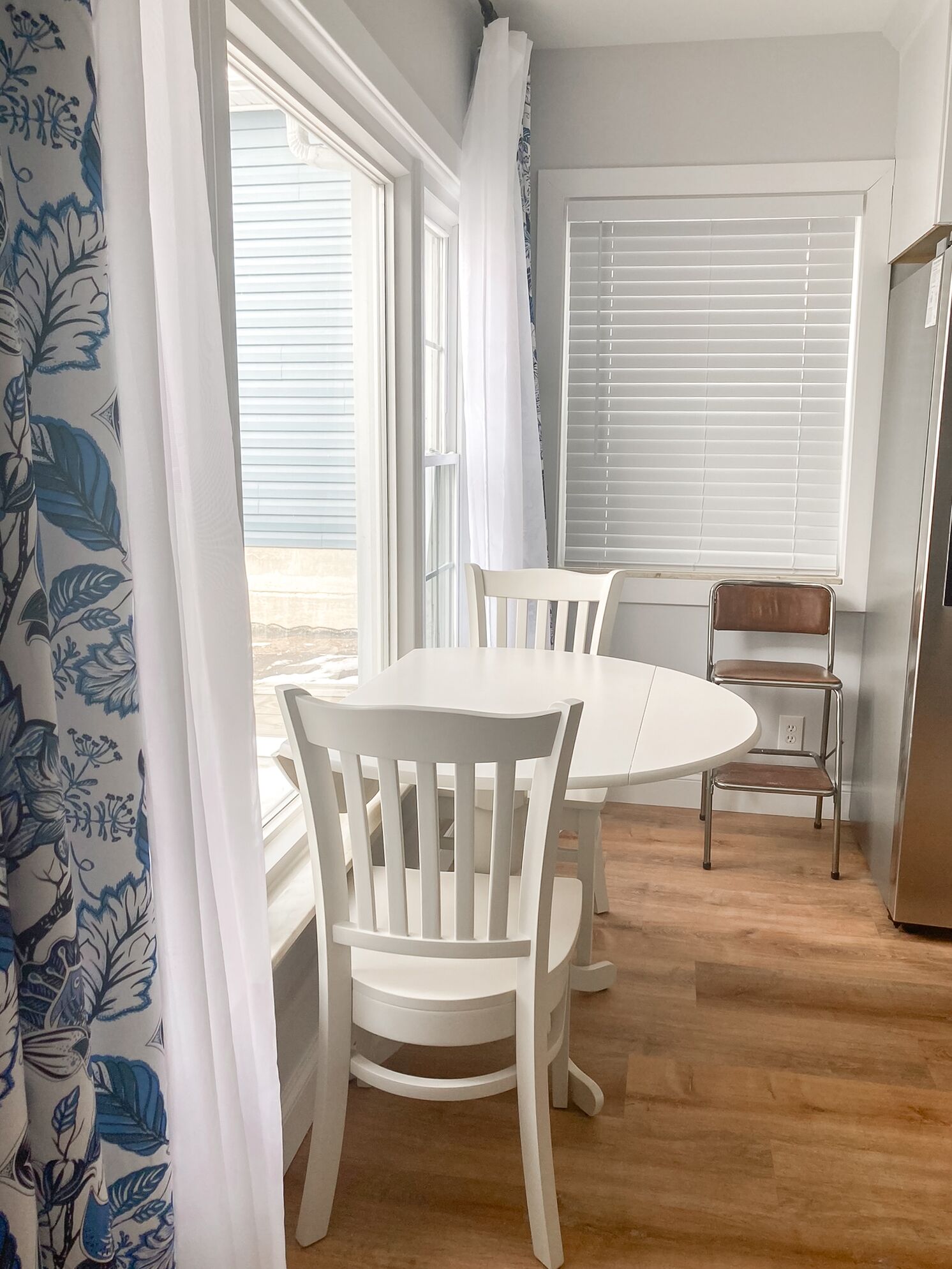 Un coin repas comprenant une table ronde et quatre chaises blanches, une chaise en métal avec un siège rembourré et un réfrigérateur.