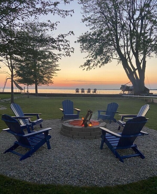A circular seating area with Adirondack chairs around a fire pit, overlooking a body of water at sunset.