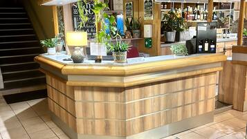 Zona de recepción con mostrador de madera, plantas en macetas, letrero de recepción y una barra con botellas y vasos.