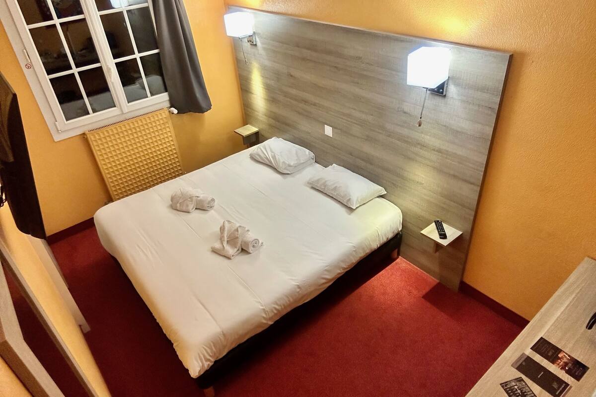 Een hotelkamer met een bed, twee bedlampjes, een raam met gordijnen, een wandheater en een klein tafeltje met een afstandsbediening.