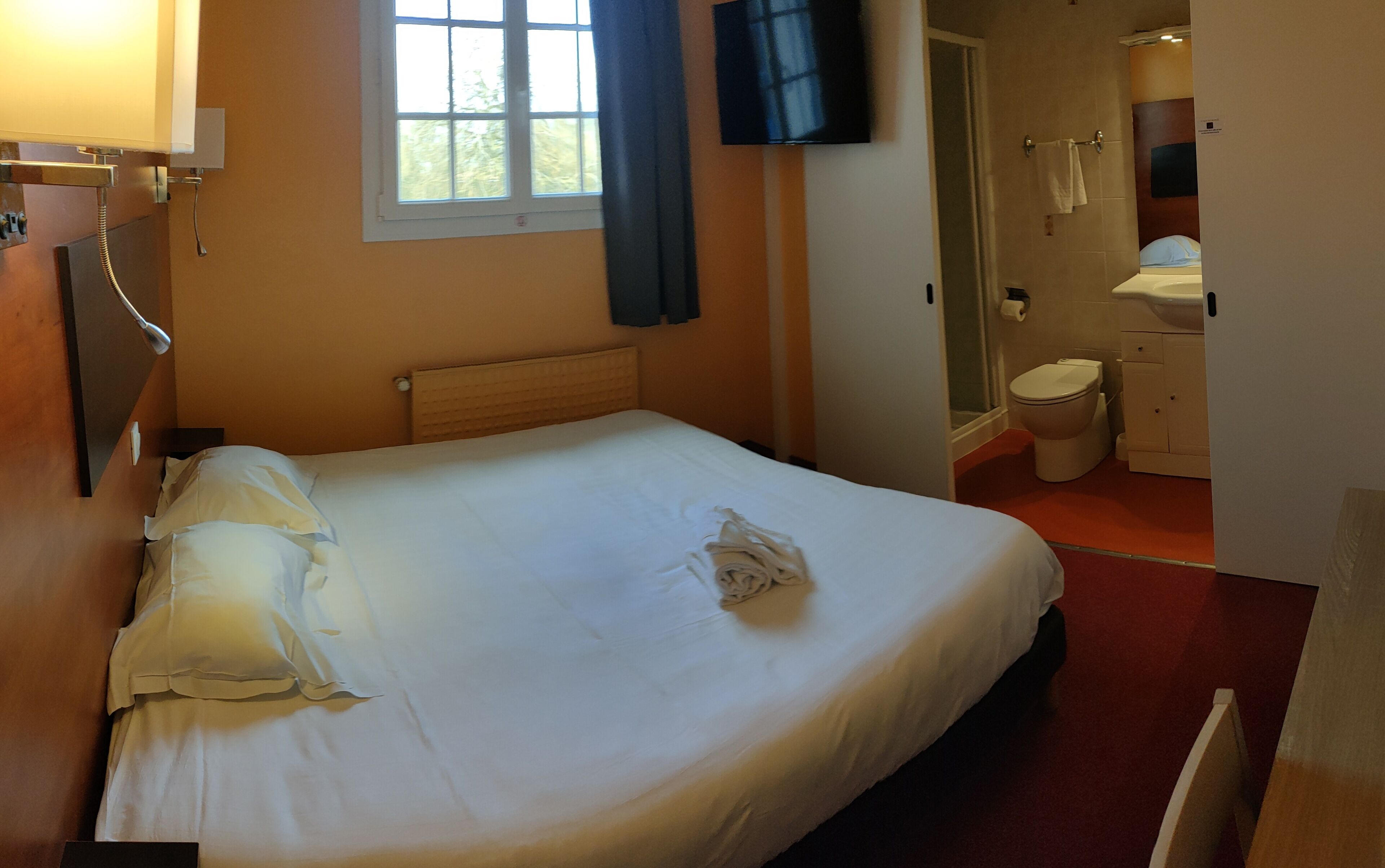 Quarto de hotel com cama, televisão, janela com cortinas, banheiro com vaso sanitário e pia, e chuveiro.