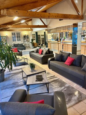 Een ruime woonkamer met een hoge plafond, een zithoek met banken en fauteuils, een salontafel, en een potplant.