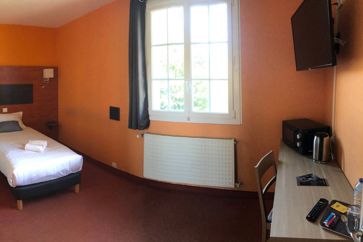 Een hotelkamer met een bed, een bureau met waterkoker, een radiator en een raam met gordijnen.
