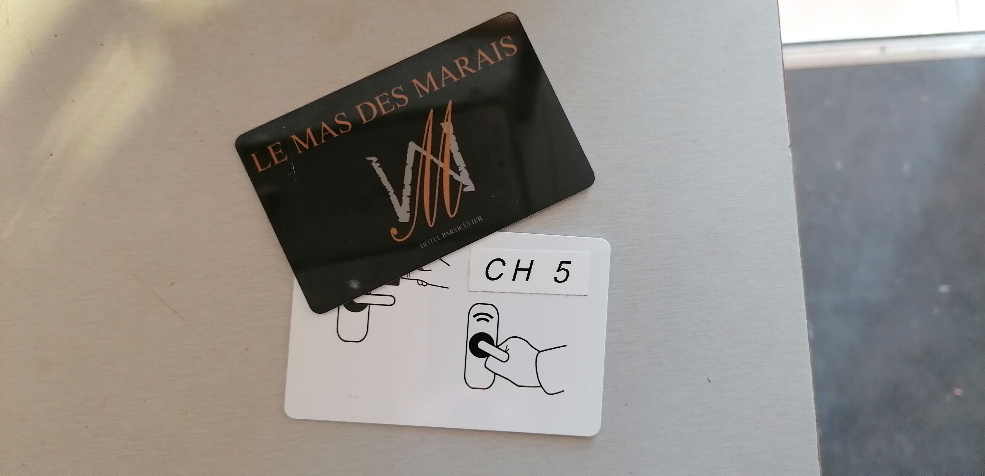 Una carpeta negra con la leyenda "LE MAS DES MARAIS" y un logotipo de hotel, junto con una tarjeta blanca que indica "CH 5" y una ilustración de una mano.