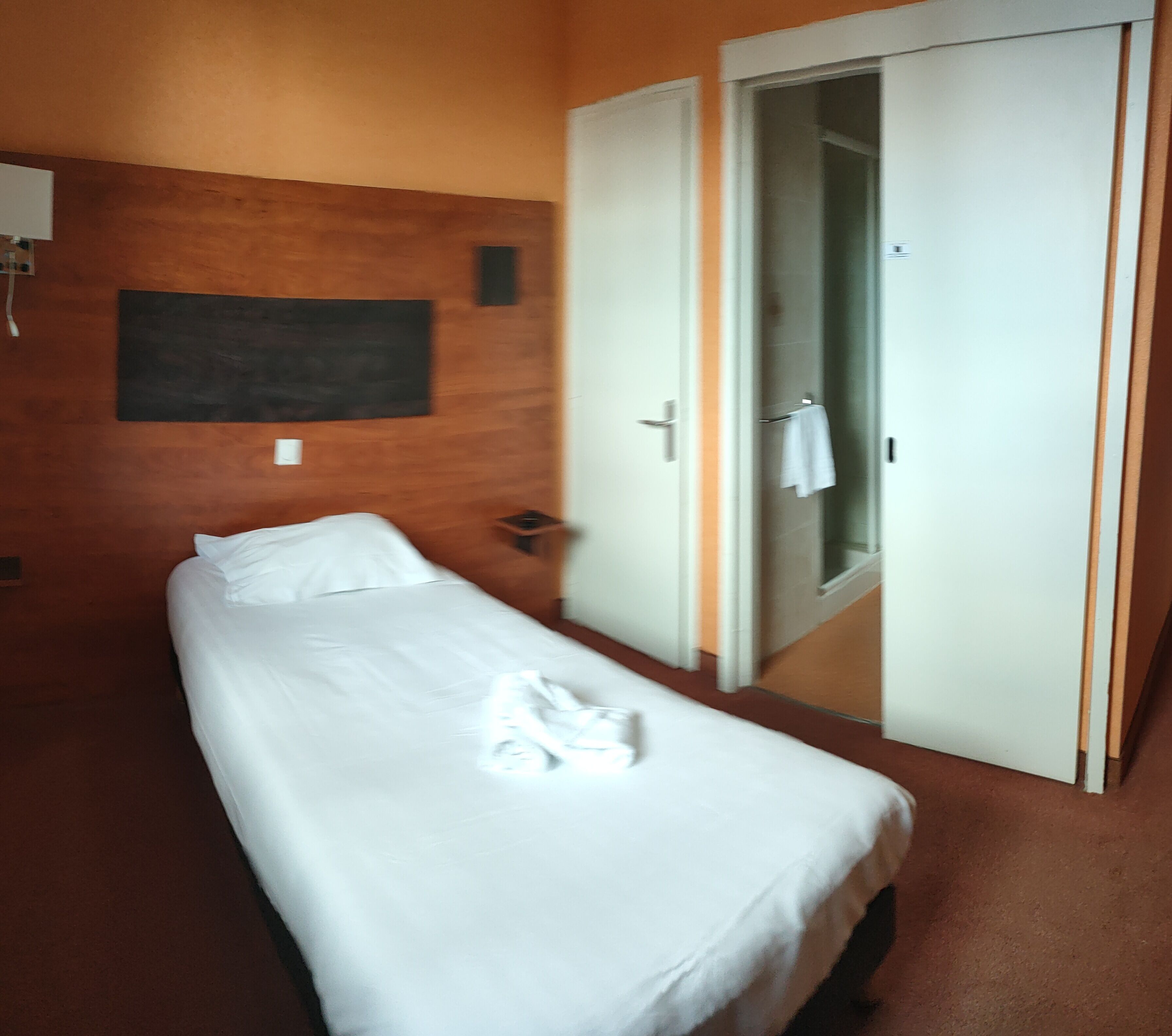 Una cama individual con ropa de cama blanca, un cabecero de madera, una mesita de noche pequeña y una toalla blanca sobre la cama.