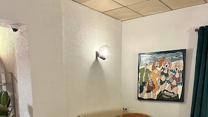 A cozy room with wicker furniture, a radiator, a potted plant, and a colorful painting on the wall. - Le Mas des Marais (Machecoul-Saint-Même)