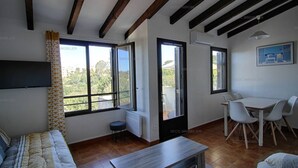 A spacious living room with a sofa, a coffee table, and a dining area with chairs. There are large windows providing a view of the outdoors.