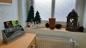Huoneessa on puinen työpöytä, patteri, pitsiverhoilla varustettu ikkuna, tervetulotoivotus, koristeellisia käpyjä ja lyhty.