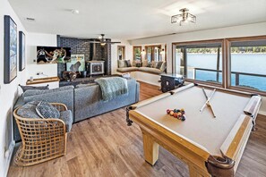 A modern living room with a pool table, a fireplace, and a large window overlooking a body of water.