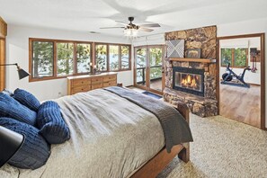 A bedroom with a bed, a fireplace, a ceiling fan, and large windows.